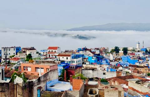       Panoramic view over the blue-painted houses of Chefchaouen with a blanket of clouds below distant mountains.
  
