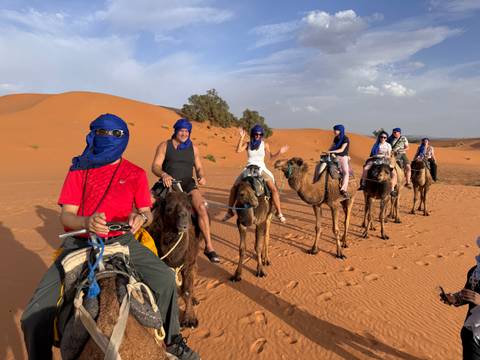       A line of travelers riding camels across orange Sahara sand dunes, led by a guide under a partly cloudy sky.
  