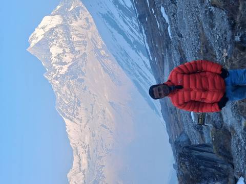       Portrait of hiker in orange jacket with dramatic snowy Himalayan backdrop in early morning light.
  