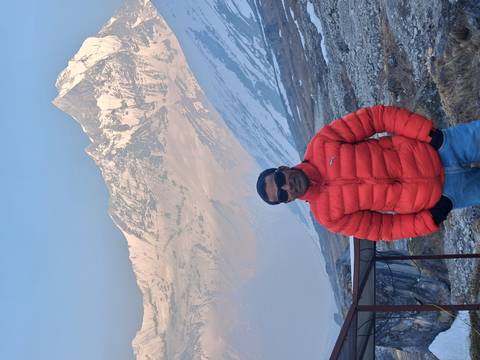       Man in a bright red down jacket posing with snow-covered Himalayan peaks towering behind him.
  