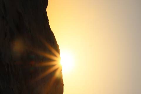       Golden sun peeks over a desert ridge casting long sunrays into the sky.
  