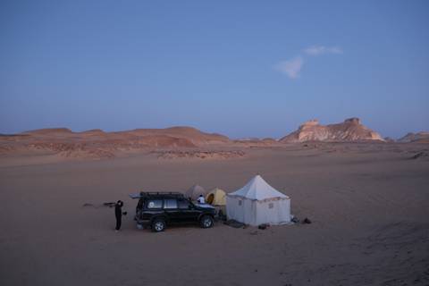       4x4 vehicle, canvas tents and solitary camper set up in vast desert at twilight.
  