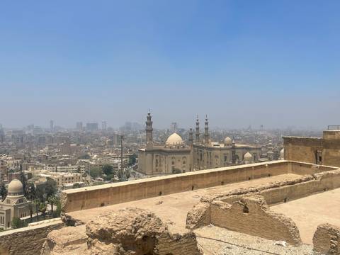      Hazy panoramic view of Cairo skyline with mosques seen from citadel walls under noon haze.
  