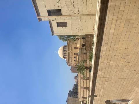       Small domed church with cross peeking over high sandstone wall beneath blue sky.
  