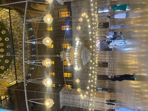       Opulent Ottoman-style mosque interior with hanging globe lamps and marble columns, tourists observing.
  