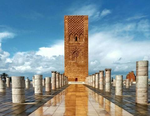       Tall sandstone tower with ornate carvings framed by rows of stubby marble columns beneath a partly cloudy sky.
  