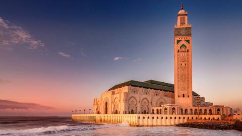       Grand coastal mosque with high minaret bathed in warm sunset light beside gentle ocean waves.
  
