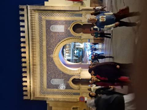       Large ornate mosaic city gate illuminated at dusk with crowds of people walking beneath its arch.
  