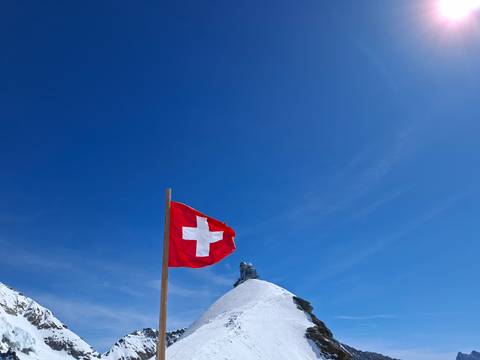       Swiss flag flying against a deep blue sky above a snowy alpine peak with viewing platform.
  