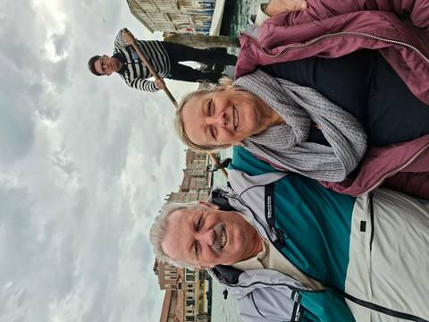       Couple sitting in a gondola with gondolier rowing through Venice’s canals under cloudy skies.
  