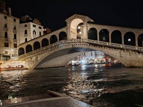       Night view of Venice’s illuminated Rialto Bridge arching over the Grand Canal with reflections on the water.
  