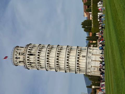       Iconic Leaning Tower of Pisa rising above green lawn with visitors and blue sky.
  