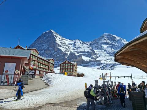      Snowy alpine village at Kleine Scheidegg with skiers, wooden lodges, and imposing Eiger north face under clear sky.
  