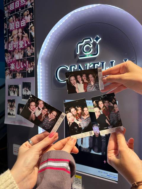       Hand holding photo-booth strips of smiling friends inside a lively Japanese arcade.
  