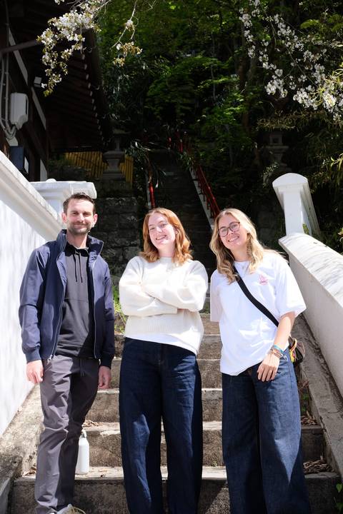       Three travellers smiling on outdoor stone steps with lush greenery and shrine railings behind.
  
