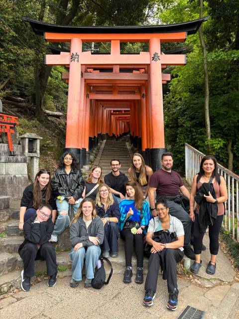      Travellers pose at the foot of a stairway lined with vermilion torii gates at Fushimi Inari Shrine.
  