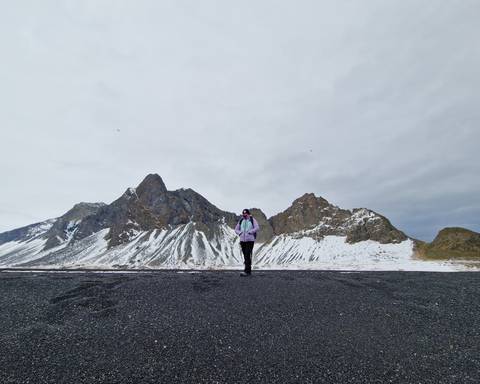       Solo hiker on a black sand plain with snow-striped jagged peaks rising behind
  