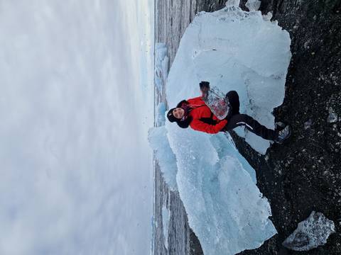       Traveler in red jacket kneeling on clear blue ice shards on a volcanic black beach
  