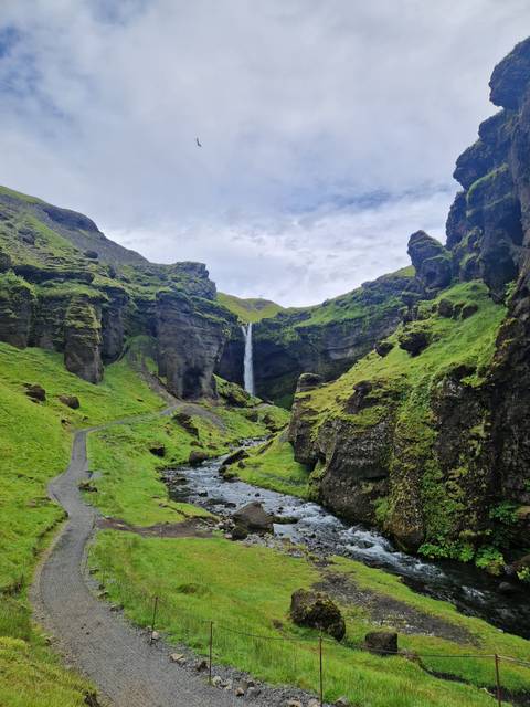       Tall slender waterfall plunging into a lush green canyon with a stream winding below
  
