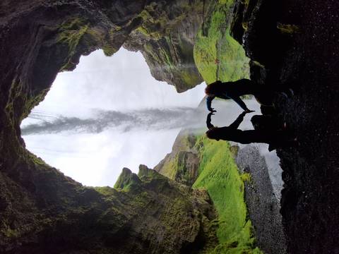       Two silhouetted travelers forming a heart shape inside a cave behind a waterfall curtain
  