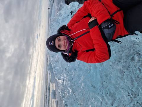       Young woman in a red jacket leans against a vivid blue ice block at a glacial beach under a cloudy sky.
  