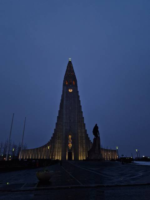       Hallgrimskirkja church tower lit up against a deep blue evening sky with a small statue silhouette in front.
  