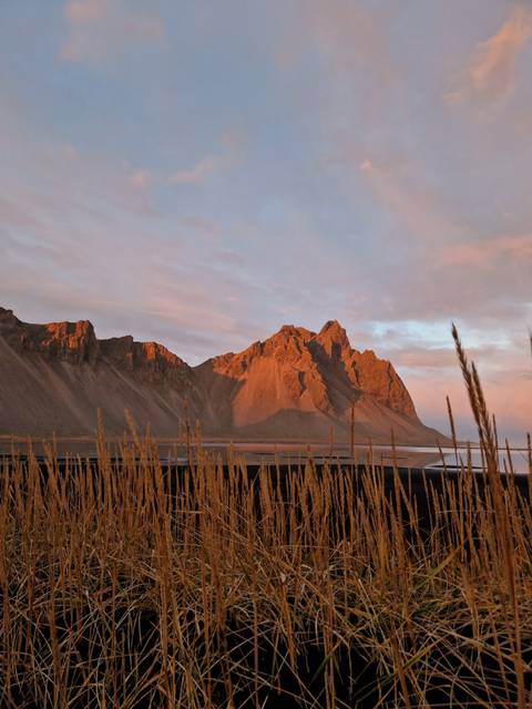       Dramatic mountain peaks glow orange in low sun, framed by tall grasses and a calm inlet.
  