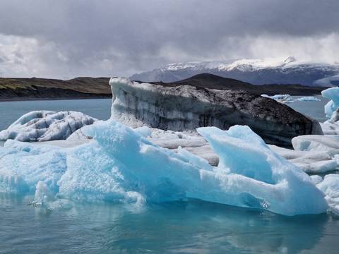       Jagged blue and black icebergs float in turquoise water beneath cloudy skies and distant snowy peaks.
  