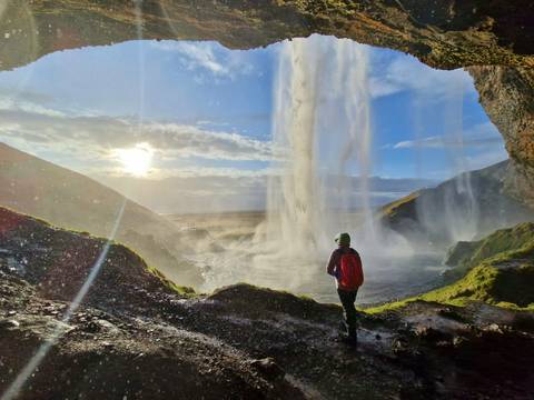       Traveler in red jacket stands inside a misty cave behind a waterfall as sunlight streams through.
  