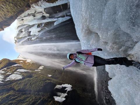       Ice-clad waterfall towers over a climber posing on frozen ground with crampons and helmet.
  