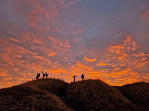      Silhouettes of friends celebrate atop grassy dunes against a dramatic fiery sunset sky.
  