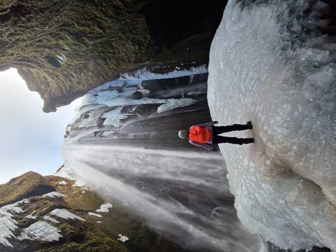       Adventurer stands on ice at the base of a towering frozen waterfall inside a steep gorge.
  