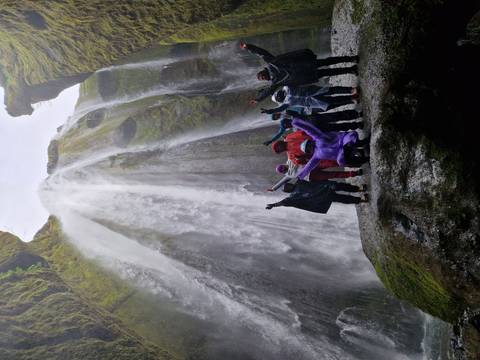       Rain-clad group raises arms in triumph beneath a thundering waterfall inside a rocky cavern.
  