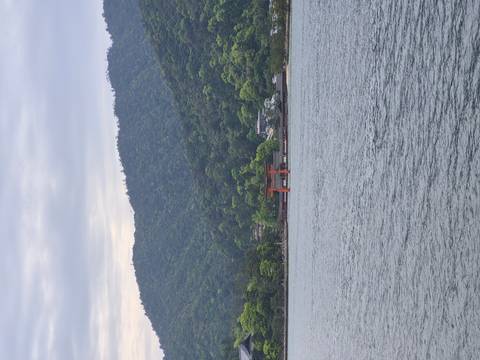       Iconic red torii gate of Miyajima stands in the water against forested mountains and cloudy sky.
  