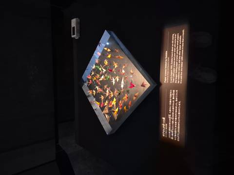       Exhibit of colorful folded paper cranes displayed under soft lighting with bilingual explanation panel.
  