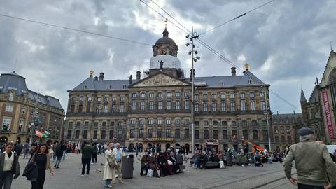       Crowds gather in Amsterdam's Dam Square in front of the Royal Palace under dramatic grey clouds.
  