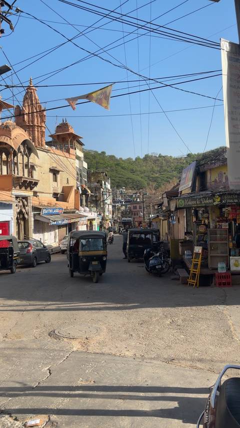       Narrow Indian town street with auto-rickshaws, shops and hills covered in trees beyond.
  