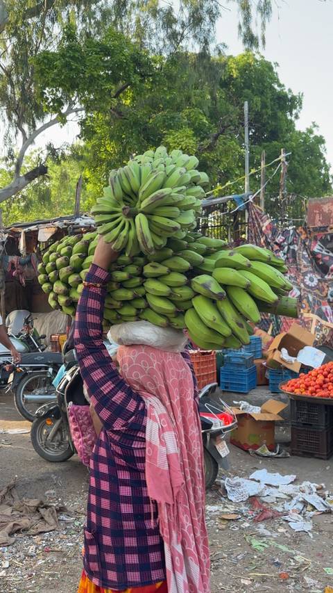       Street vendor balances a huge bunch of green bananas on their head in a busy market.
  
