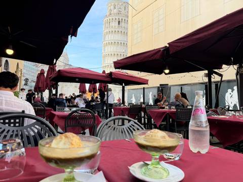       Outdoor café with desserts and mineral water on the table, Leaning Tower of Pisa visible between burgundy umbrellas.
  