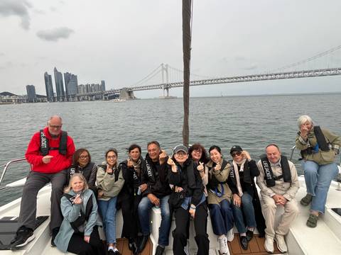      Tour group in lifejackets poses on boat deck with Busan skyline and suspension bridge behind.
  