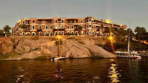       Historic riverside hotel atop granite rocks illuminated at dusk along the Nile.
  