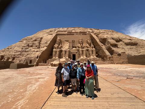       Tour group posing on a wooden walkway before the monumental rock-cut façade of Abu Simbel’s Great Temple under a clear blue sky.
  