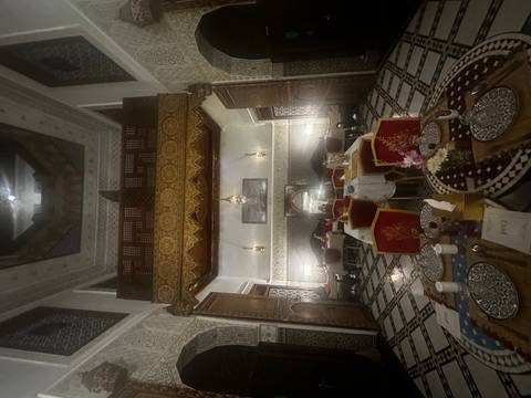       Dimly lit traditional riad dining room with patterned tiles and ornate wooden balcony.
  