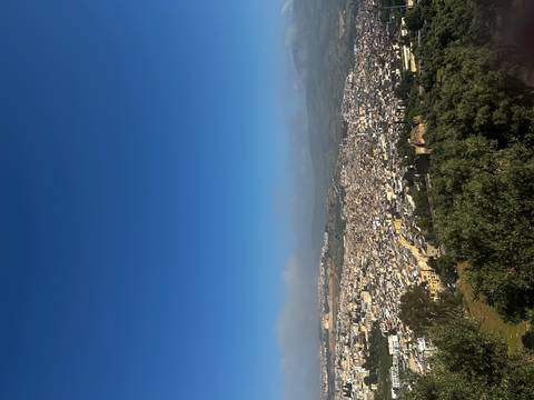      Wide panoramic view over a sprawling Moroccan city nestled between green hills under a clear blue sky.
  