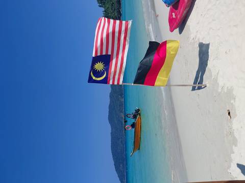       Pristine white-sand beach with Malaysian and German flags waving beside a small wooden boat on turquoise water.
  