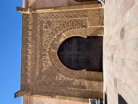       Intricately carved sandstone gateway with large arched wooden doors against a clear blue sky
  