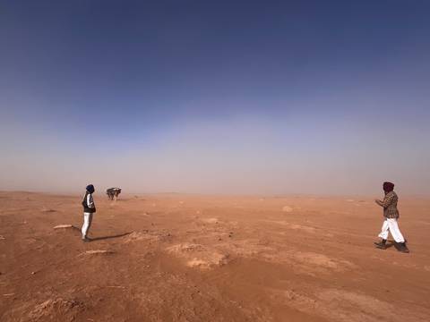       Vast dusty desert landscape with two travelers standing far apart under a clear blue sky.
  