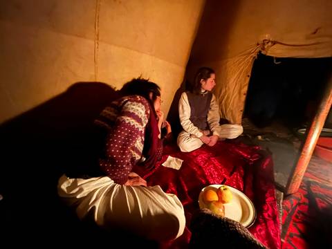       Dimly lit interior of a desert tent with two travelers sitting on red carpets beside a plate of fruit.
  