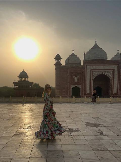       Soft sunrise light over ornate red sandstone mosque next to the Taj Mahal complex with visitors in foreground.
  