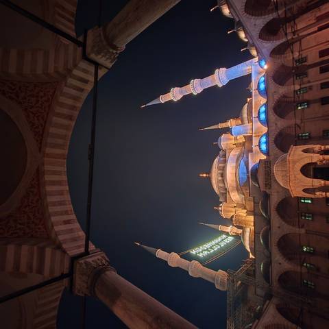       The illuminated domes and minarets of Istanbul’s Blue Mosque glow against a deep night sky, framed by an arched entrance.
  
